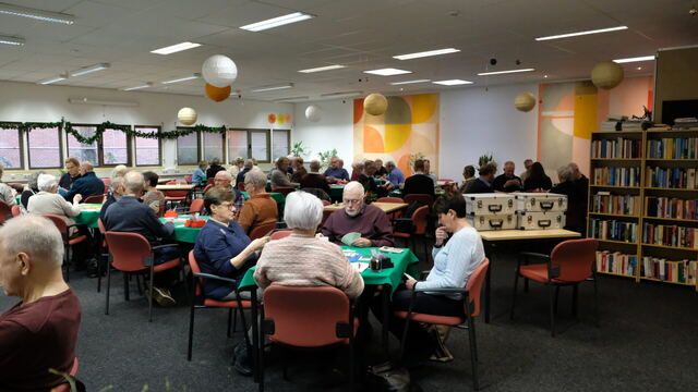 Volle zaal met spelers aan groene speeltafels tijdens het inloopcafé